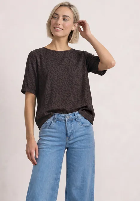 Frau trägt schwarzes T-Shirt mit Muster und blaue Jeans vor hellem Hintergrund.