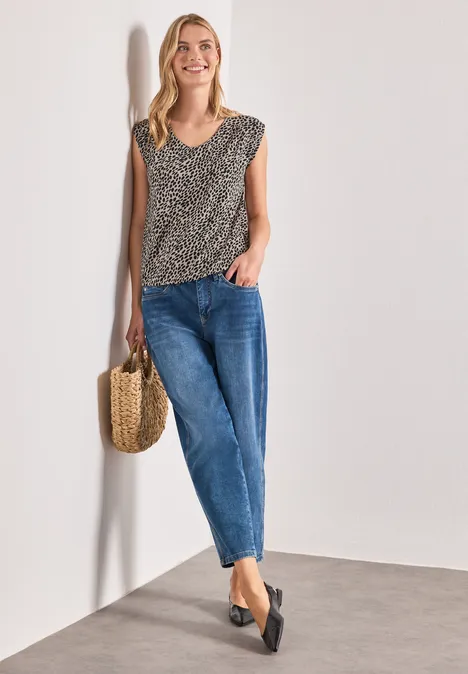 Frau lehnt an Wand, trägt Leo-Top, blaue Jeans, schwarze Ballerinas und hält eine Strohtasche.