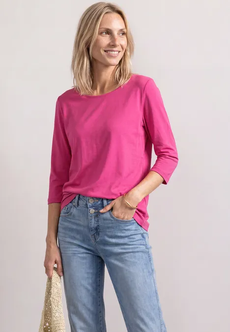 Eine lächelnde Frau in pinkem 3/4-Arm-Shirt und Jeans hält eine geflochtene Tasche.
