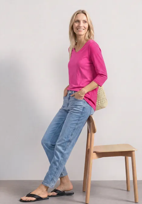 Frau in pinkem Shirt, Jeans und schwarzen Sandalen lehnt lässig an einem Hocker vor weißem Hintergrund.