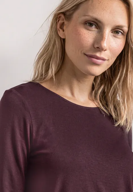 3/4-Arm Shirt mit U-Boot-Ausschnitt rustic rouge Frau mit blonden Haaren und dezentem Lächeln in einem dunkelvioletten Langarmshirt vor hellem Hintergrund.