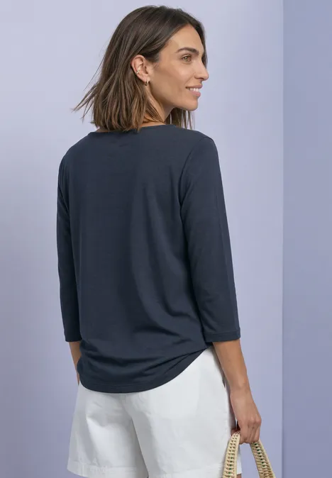 Frau in dunkelgrauem 3/4-Arm-Shirt und weißen Shorts, seitlich gewandt mit Korbtasche in Hand vor lila Hintergrund.