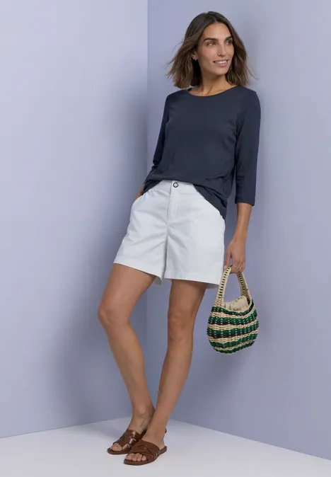 Frau in weißer Shorts, dunkelblauem Oberteil, Sandalen, hält einen gehäkelten Korb vor pastellfarbener Wand.