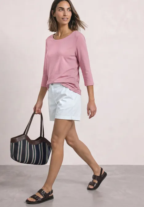 3/4-Arm Shirt mit U-Boot-Ausschnitt vintage blush Frau in rosa Shirt und weißen Shorts mit gestreifter Tasche und schwarzen Sandalen vor hellem Hintergrund.