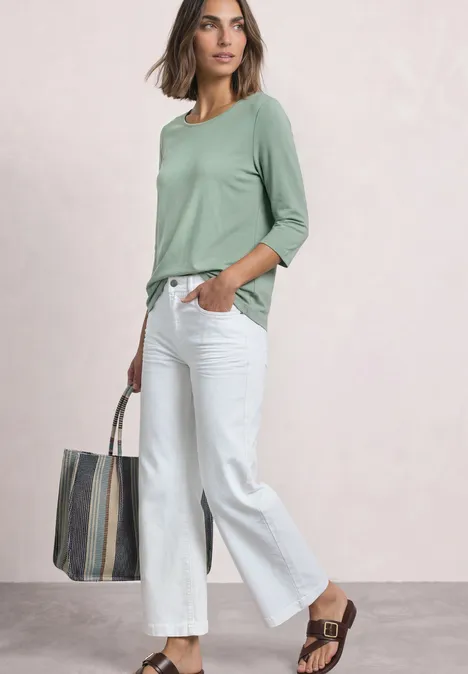 3/4-Arm Shirt mit U-Boot-Ausschnitt eucalyptus green Eine Frau in grüner Bluse, weißer Hose und Tasche steht entspannt.