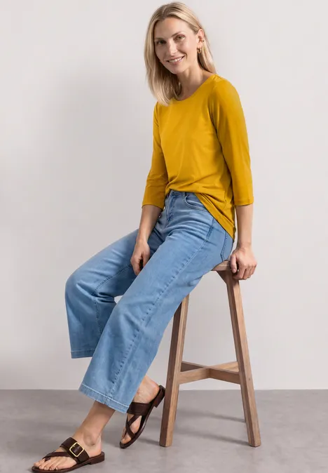 3/4-Arm Shirt mit U-Boot-Ausschnitt saffron yellow Frau sitzt auf Hocker, trägt senfgelben 3/4-Arm-Pullover, weite Jeans und schwarze Sandalen.