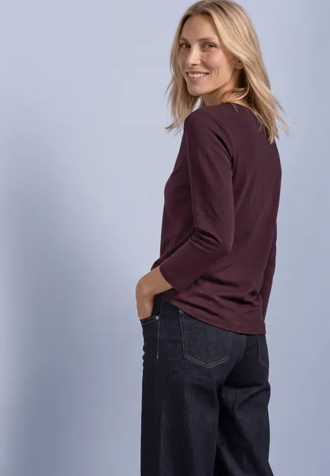 Eine Frau in violettem Langarmshirt und dunklen Jeans blickt über ihre Schulter vor hellem Hintergrund.