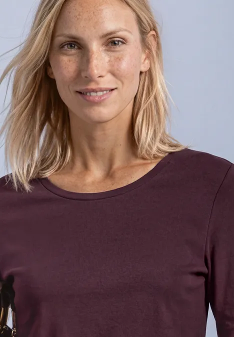 Lächelnde Frau mit blonden Haaren und Sommersprossen in einem langärmeligen, dunkelvioletten Shirt.