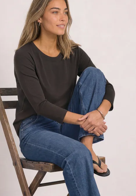 Frau in schwarzem Oberteil und blauen Jeans sitzt lächelnd seitlich auf einer Holzleiter.