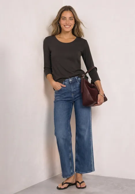 Frau in schwarzem Shirt und weitem Jeans, Sandalen und brauner Tasche, lächelt vor hellem Hintergrund.