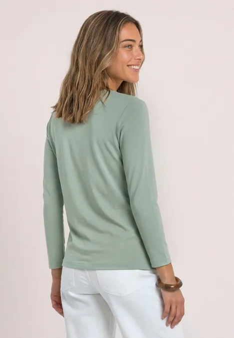 Frau im grünen Langarmshirt und weißer Hose, schaut seitlich lächelnd.