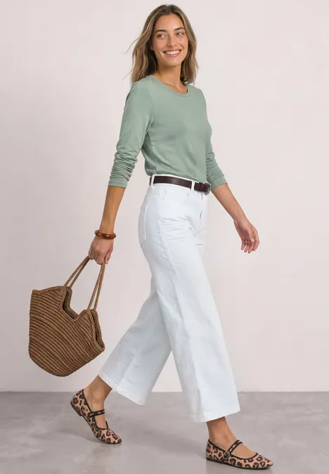 Eine Frau trägt weiße Culottes, grünes Oberteil, Korbtasche und Sandalen, während sie lächelnd spaziert.