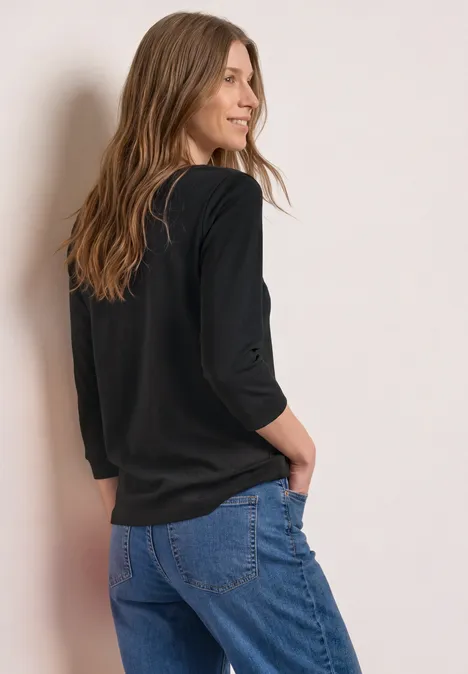 Eine Frau von hinten in schwarzem Oberteil und blauen Jeans, die entspannt an einer Wand steht.