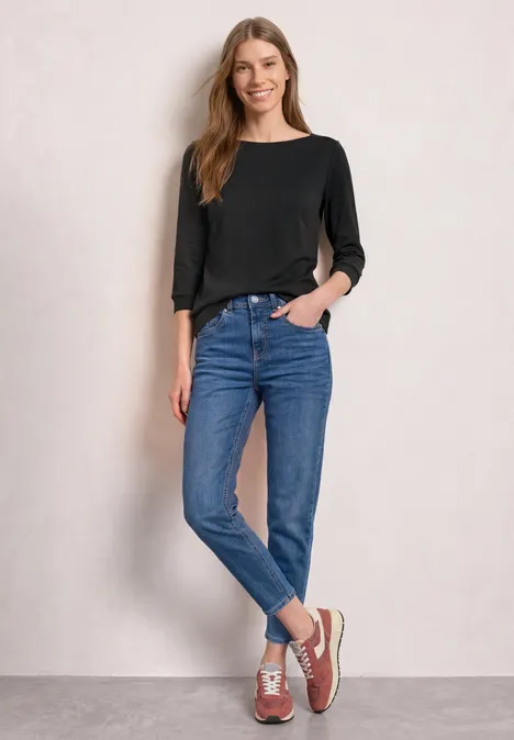 Eine Frau in schwarzem Langarmshirt, blauen Jeans und Sneakers steht lässig vor einer hellen Wand.