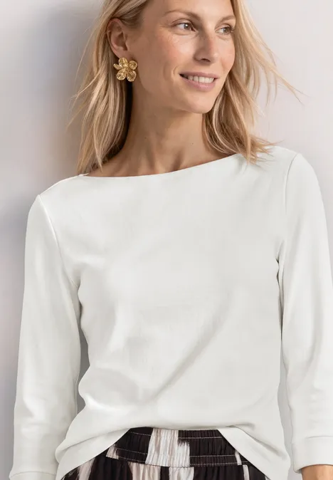 Frau trägt weiße 3/4-Ärmel-Bluse mit U-Boot-Ausschnitt, gemusterten Rock und Blumenohrring.