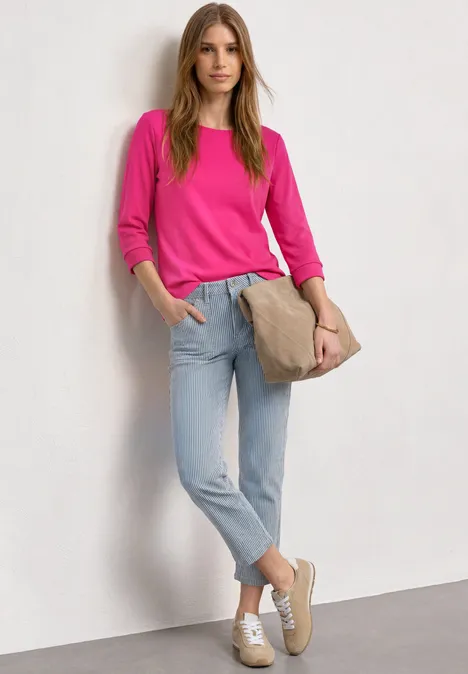 Junge Frau lehnt an weißer Wand, trägt pinkes Shirt, helle Jeans, weiße Sneaker und eine braune Umhängetasche.