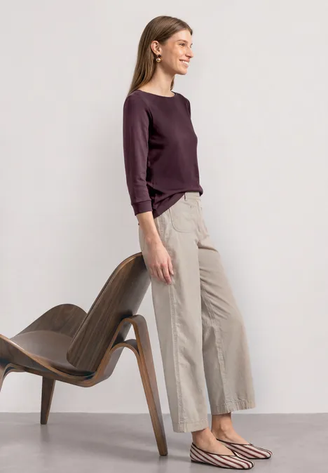 Eine Frau im seitlichen Profil, trägt lila Pullover und weite beige Hosen, steht neben einem modernen Holzstuhl.