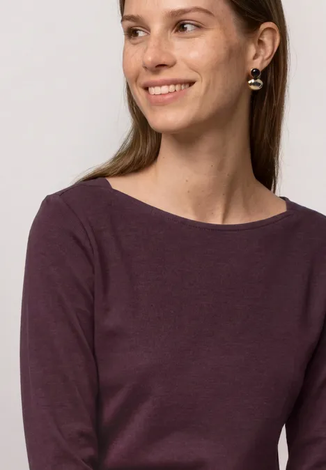 Frau mit hellem Haar trägt ein dunkelviolettes Langarmshirt, Gesicht unscharf, Ohr mit Ohrring sichtbar.