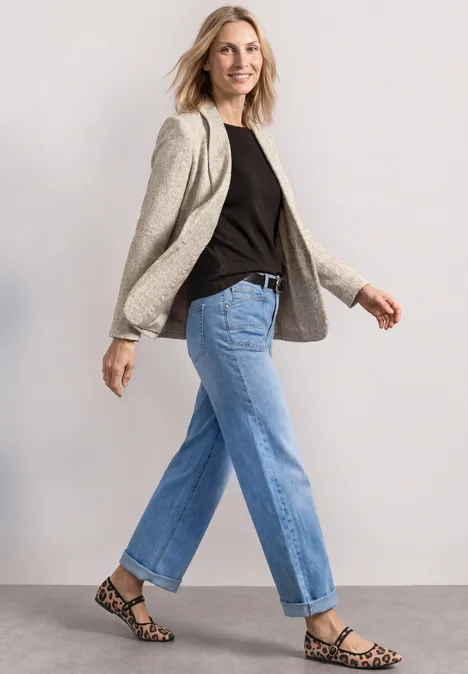 Frau in beigem Blazer, schwarzem Shirt, blauen Jeans und Leoparden-Sandalen geht nach rechts.