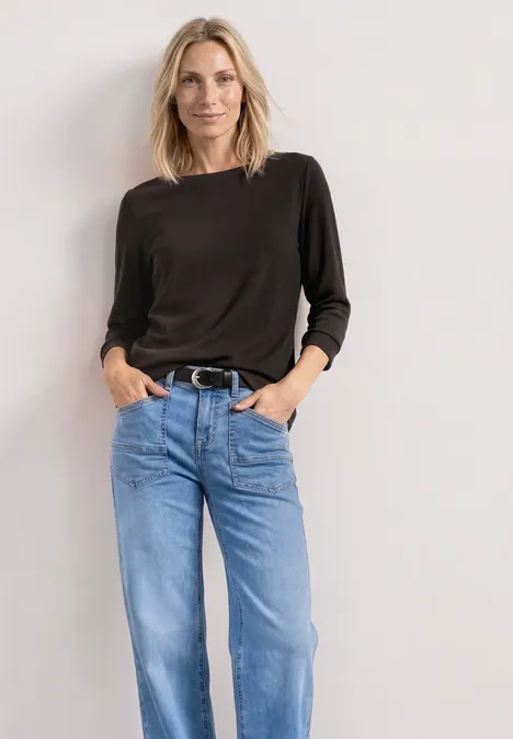 Person vor heller Wand in schwarzem Langarmshirt und blauen Jeans mit Gürtel.