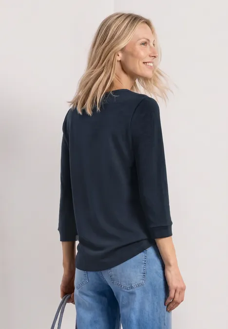 Frau von hinten trägt dunkelblauen Pullover und blaue Jeans, steht vor hellem Hintergrund.