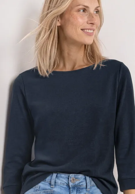Eine Frau mit schulterlangem blondem Haar trägt ein schlichtes dunkelblaues 3/4-Ärmel-Shirt vor hellem Hintergrund