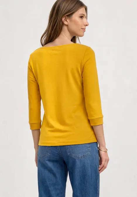 Eine Frau im Profil von hinten, trägt ein senfgelbes 3/4-ärmeliges Oberteil und blaue Jeans.