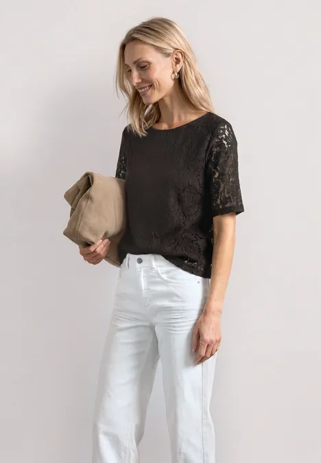 Frau mit schwarzer Spitzenbluse, weißen Jeans, hält einen beigen Hut lächelnd in der Hand.