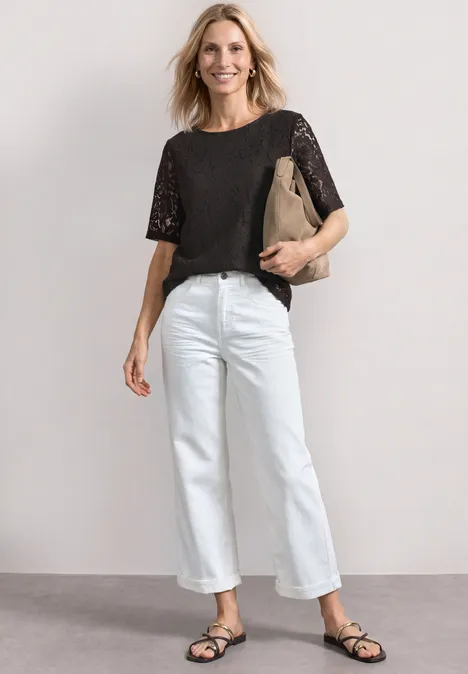 Frau in schwarzer Spitzenbluse, weißer Jeans, schwarzen Sandalen, lächelnd mit beiger Clutch vor neutralem Hintergrund.
