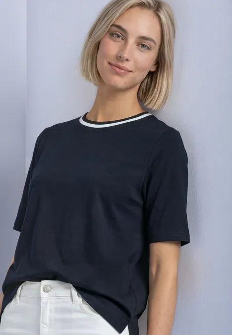 Kobieta z krótkimi blond włosami ma na sobie czarną koszulkę i białe dżinsy na jasnym tle.