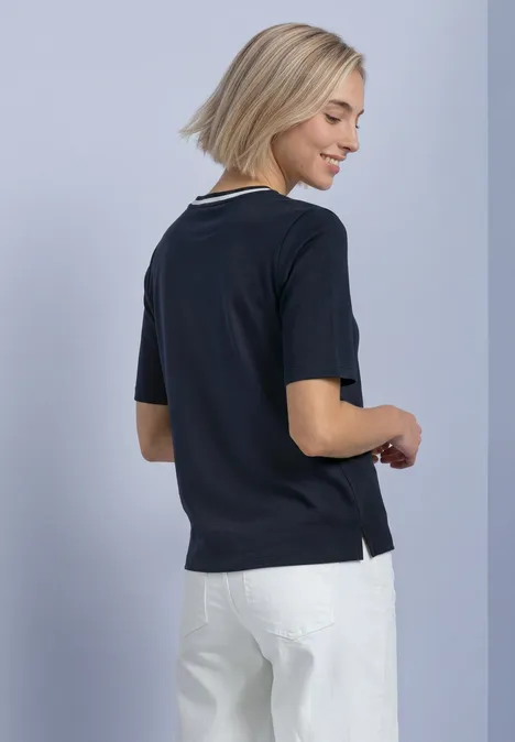 Frau im Profil von hinten, trägt dunkelblaues Shirt und weiße Hose, kurze blonde Haare, lächelt.