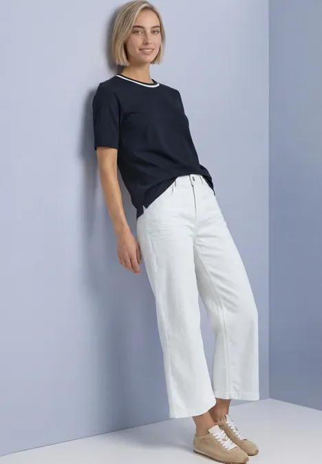 Eine blonde Frau im Bob-Look lehnt an hellblauer Wand, trägt schwarzes T-Shirt, weiße Culottes und beige Schuhe.