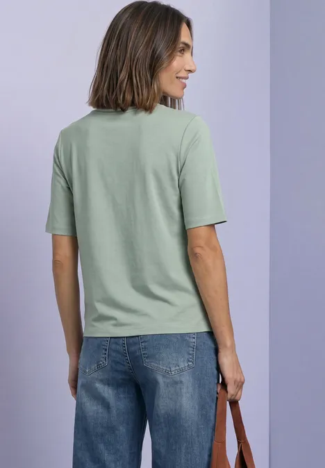 Frau von hinten in grünem T-Shirt und blauer Jeans, hält Tasche vor lila Wand.