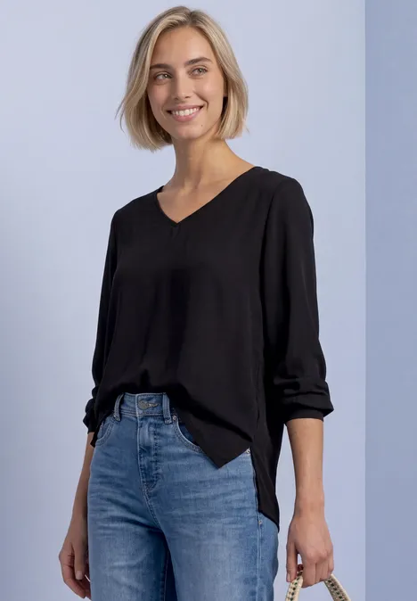 Frau mit schwarzer Bluse und blauen Jeans, lächelnd vor hellgrauem Hintergrund.