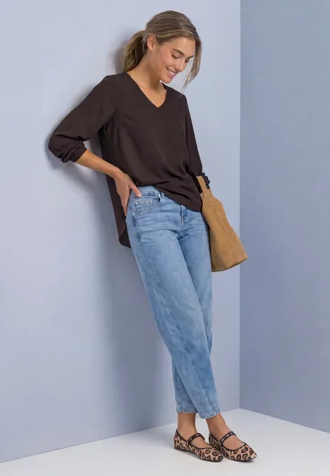 Junge Frau lehnt an heller Wand, trägt schwarzen V-Ausschnittpullover, helle Jeans, Leo-Sandalen und Schultertasche.
