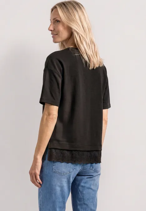 Blonde Frau von hinten in schwarzem T-Shirt mit Spitzenbesatz und blauen Jeans.