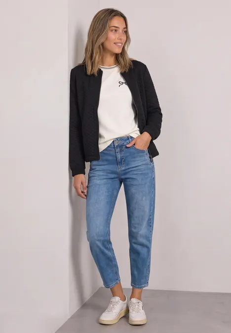 Frau in schwarzem Blazer, weißem T-Shirt, blauen Jeans und weißen Sneakern lehnt locker an der Wand.