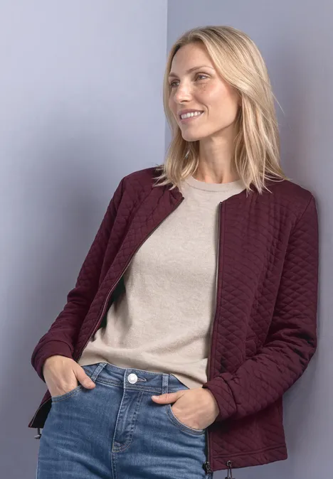 Blonde Frau in bordeauxroter Steppjacke, hellem Shirt und Jeans, seitlich lächelnd.