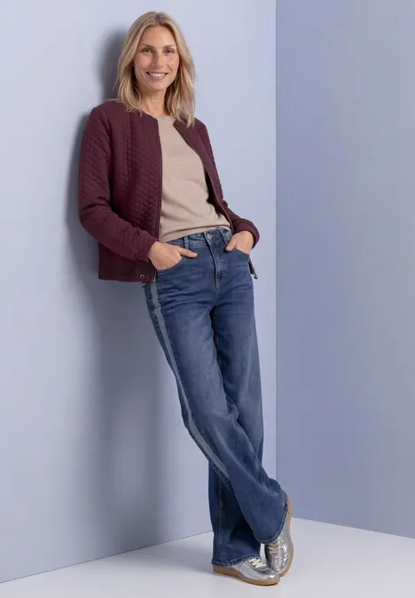 Eine Frau in Jeans, beiger Bluse und bordeauxroter Strickjacke lehnt lässig an einer hellen Wand.