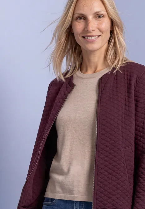 Lächelnde Frau mit blonden Haaren trägt burgunderfarbene Jacke über beigem Pullover vor pastellfarbenem Hintergrund.