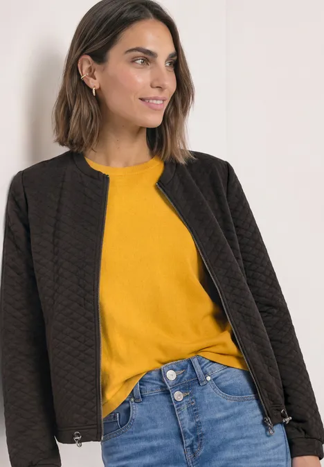 Eine Frau trägt eine gesteppt schwarze Jacke, ein gelbes Oberteil und blaue Jeans, leicht lächelnd.