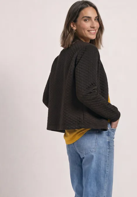 Frau in schwarzer, strukturierter Jacke, gelbem Shirt und Jeans blickt lächelnd über die Schulter.