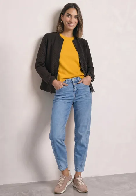 Frau trägt gelbes Oberteil, schwarze Jacke, blaue Jeans, beige Sneaker. Steht lächelnd vor neutraler Wand.