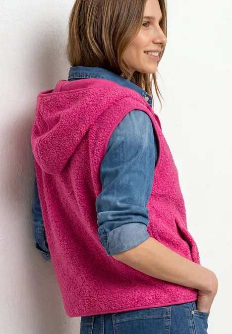 Frau in pinker, flauschiger Hoodie-Weste über Jeanshemd, seitlich stehend und lächelnd.