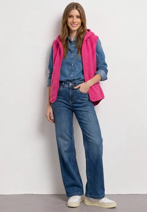 Frau in Jeanshemd und -hose, pinker Steppweste und weißen Sneakern, mit Händen in den Taschen.