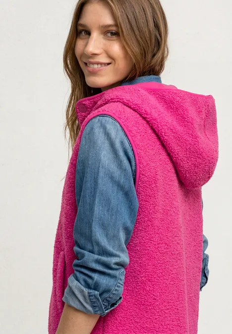 Frau in pinker Fleece-Weste mit Kapuze über Jeanshemd, lächelnd über die Schulter blickend.