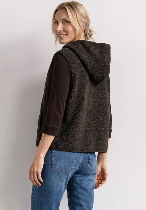 Lächelnde Frau mit blondem Haar in schwarzer Kapuzenweste, schwarzem Langarmshirt und blauer Jeans.