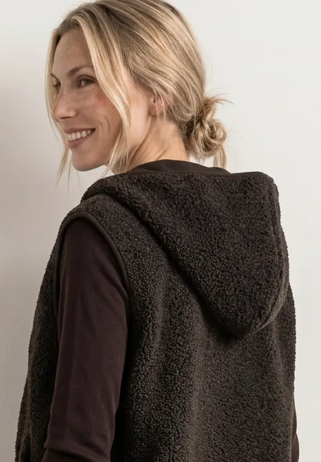 Eine lächelnde Frau mit blondem Zopf trägt eine flauschige braune Kapuzenweste über schwarzem Shirt.