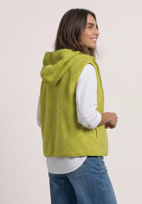 Eine Frau trägt eine grüne, kuschelige Kapuzenweste über weißem Hemd und Jeans.