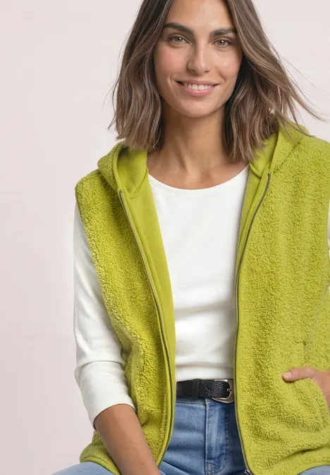 Gilet en peluche à capuche et fermeture zip apple lime Frau mit grüner Fleece-Weste mit Kapuze, weißem Shirt und blauer Jeans lächelt leicht.